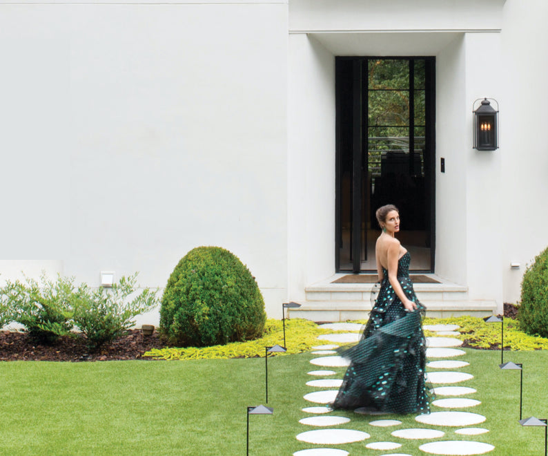A woman in an elegant green dress standing on the cobblestone walkway leading to a white house with black trim.